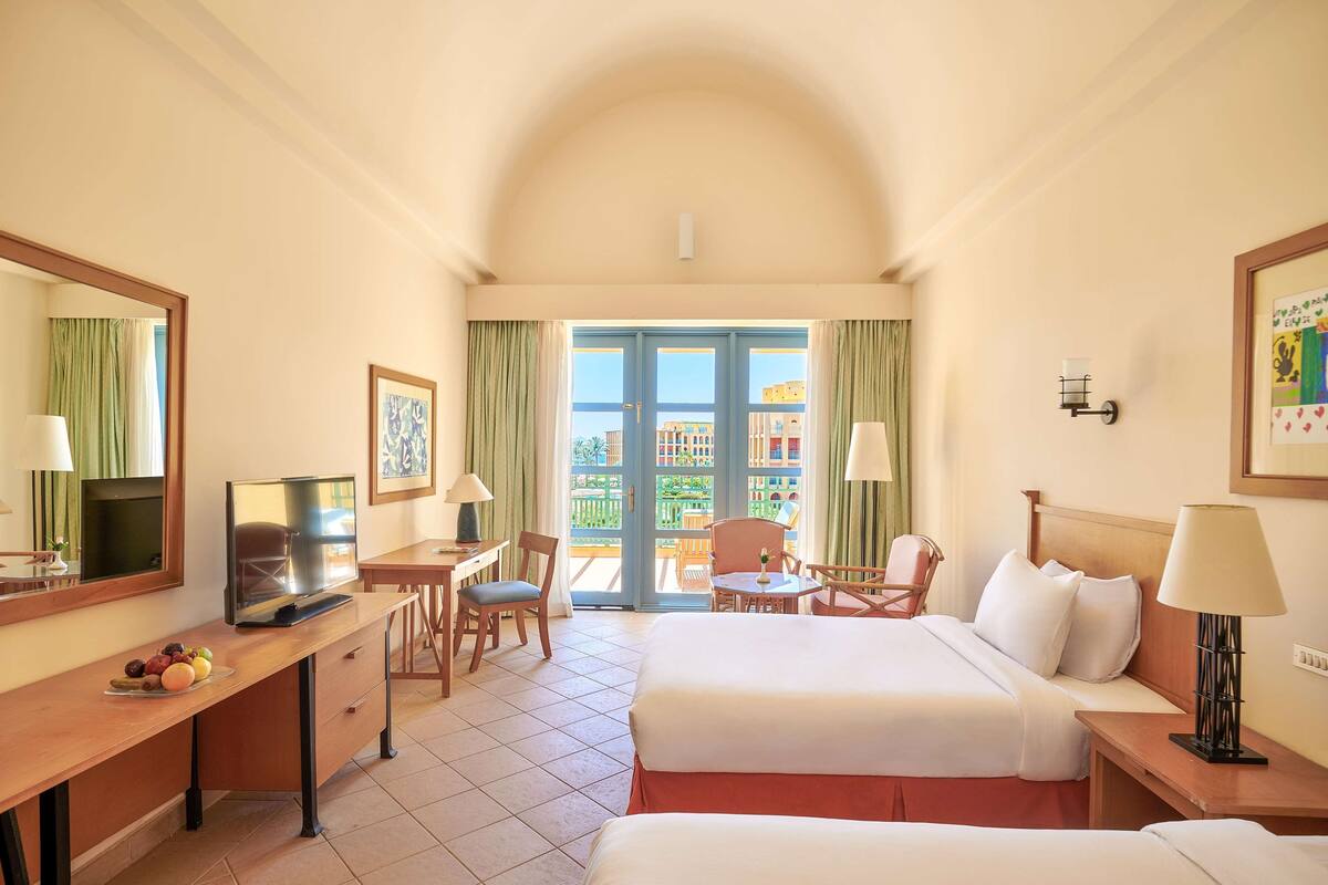 Habitación de hotel con cama, escritorio, silla, televisor y balcón con vistas.