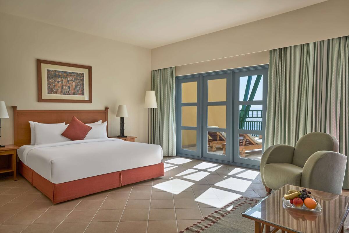 Habitación de hotel con cama, lámparas de noche, una silla, una mesa con fruta y vistas al exterior mediante una puerta corredera.