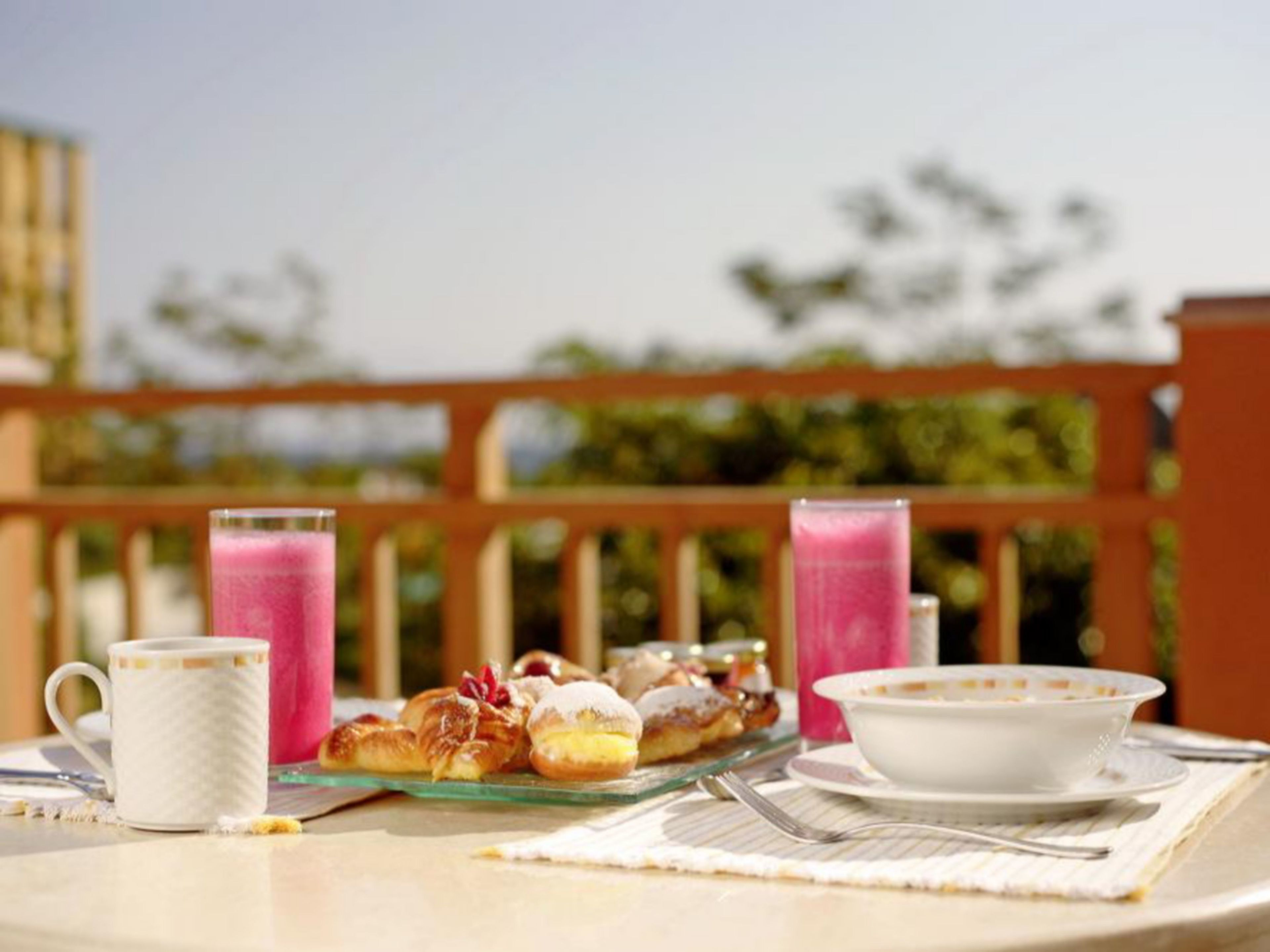 Une table avec des pâtisseries, une tasse, un bol et deux verres contenant une boisson rose, le tout disposé sur une terrasse dotée d’une balustrade en bois et avec de la verdure en arrière-plan.