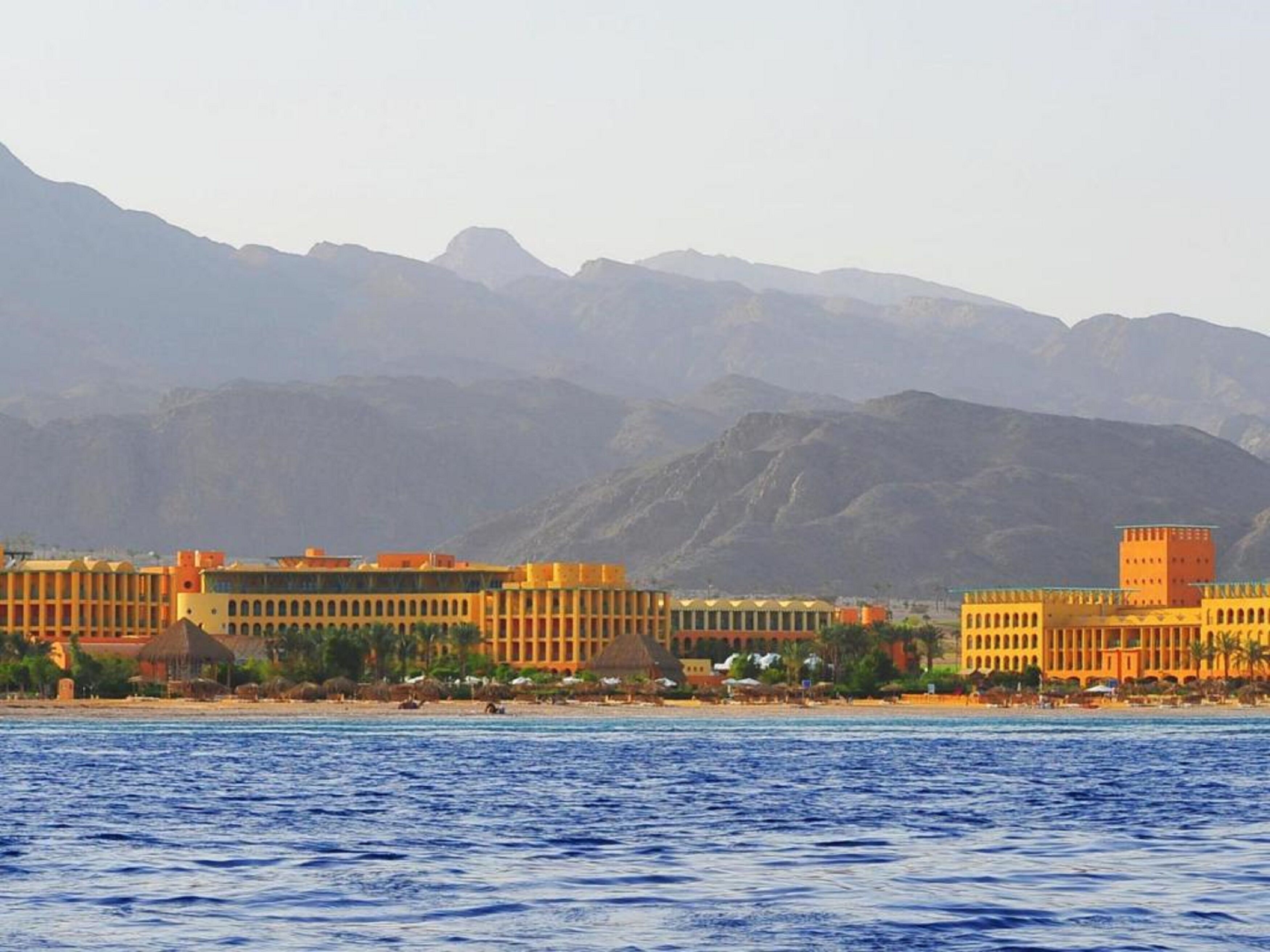 Un hotel frente al mar con varias edificaciones, una playa y montañas al fondo.