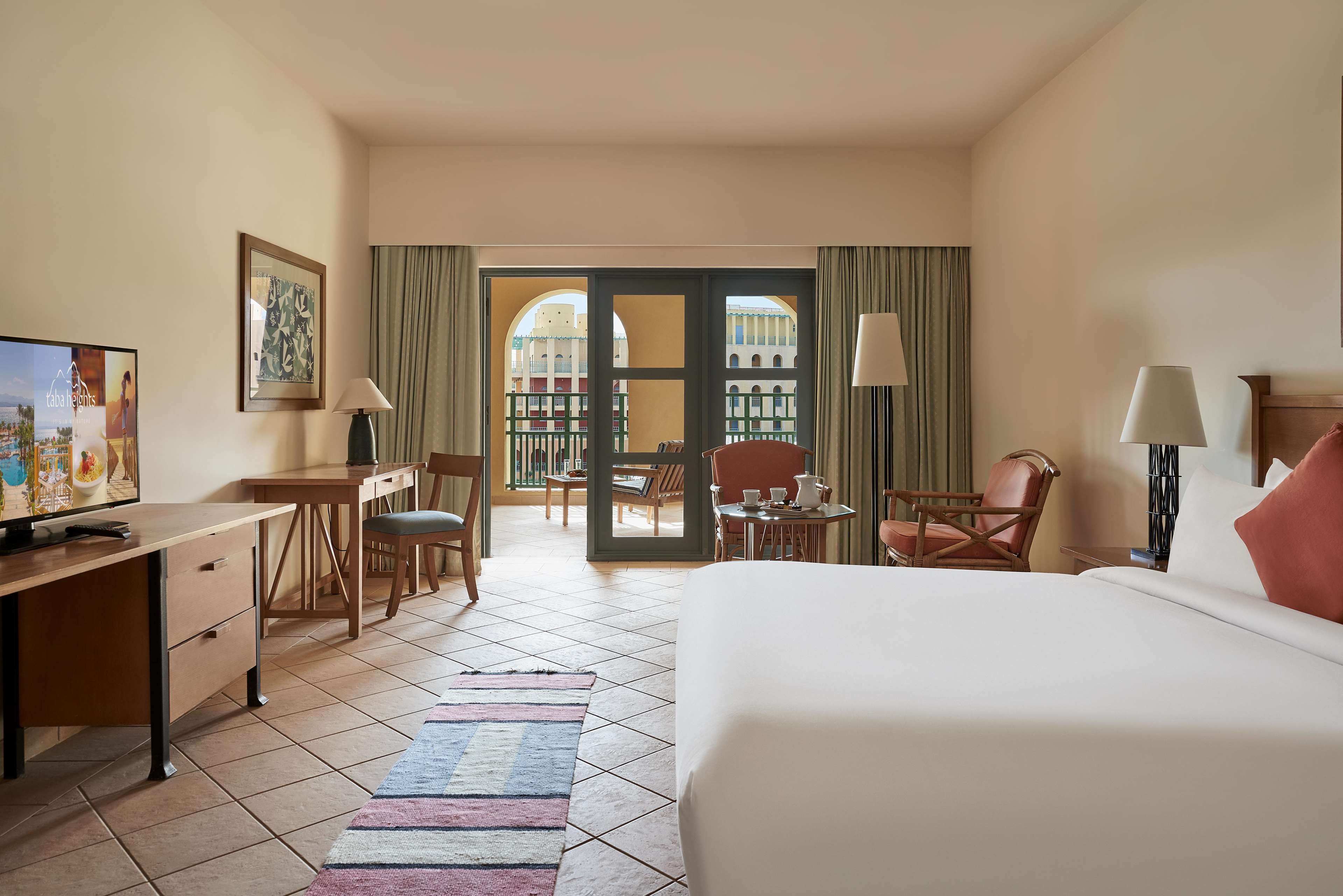 Ein Hotelzimmer mit Bett, Schreibtisch, Stühlen, einem Fernseher und Blick auf einen Außenbereich mit Balkon.