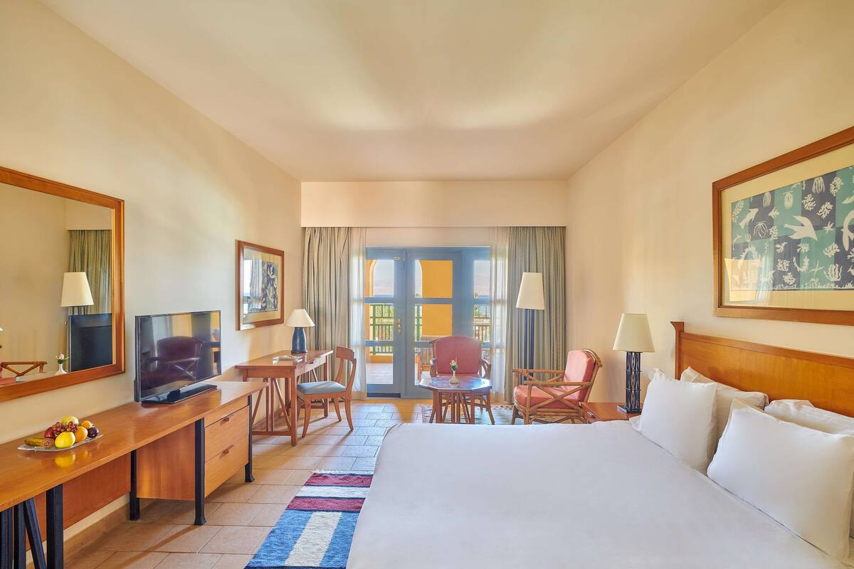 Habitación de hotel con cama, escritorio con televisor, silla, mesita y ventana con cortinas.