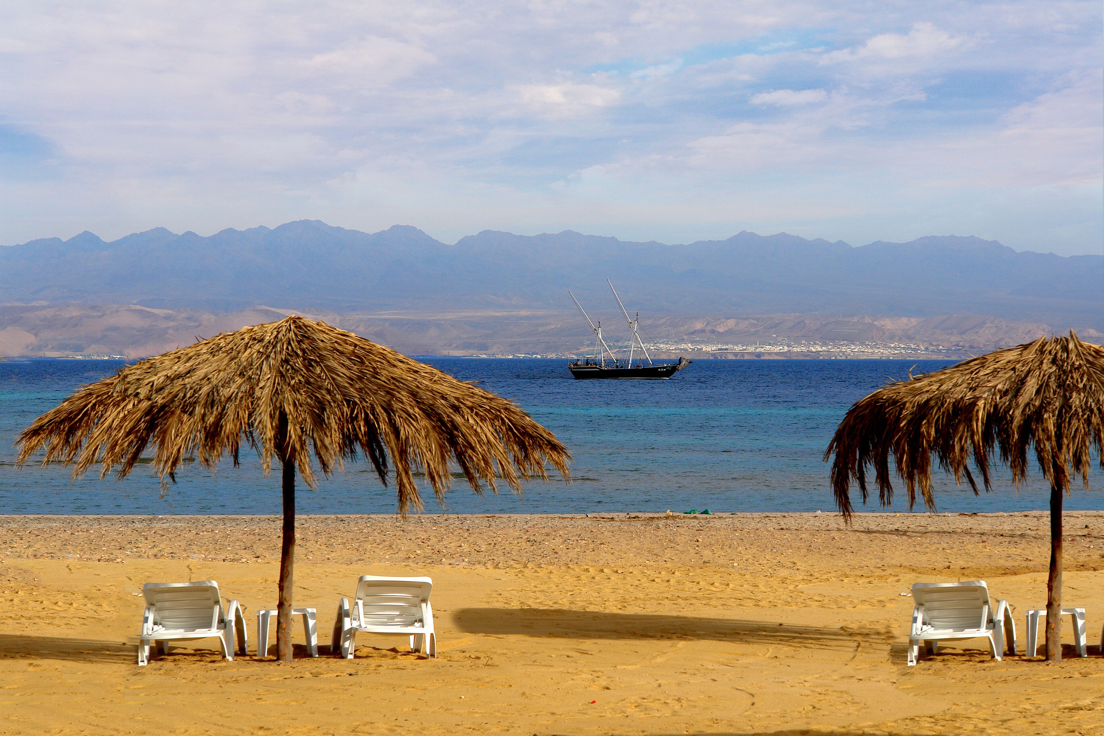 A beach with two thatched umbrellas and white lounge chairs.