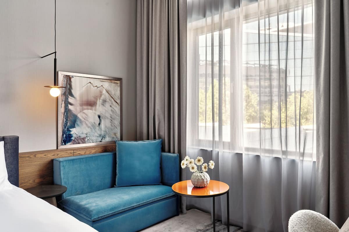 A modern hotel room with a blue sofa, a round table with a vase of flowers, a painting on the wall, and a large window with curtains.