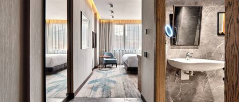 Habitación de hotel con cama, una silla, baño con lavamanos y espejo, y pasillo con piso alfombrado.