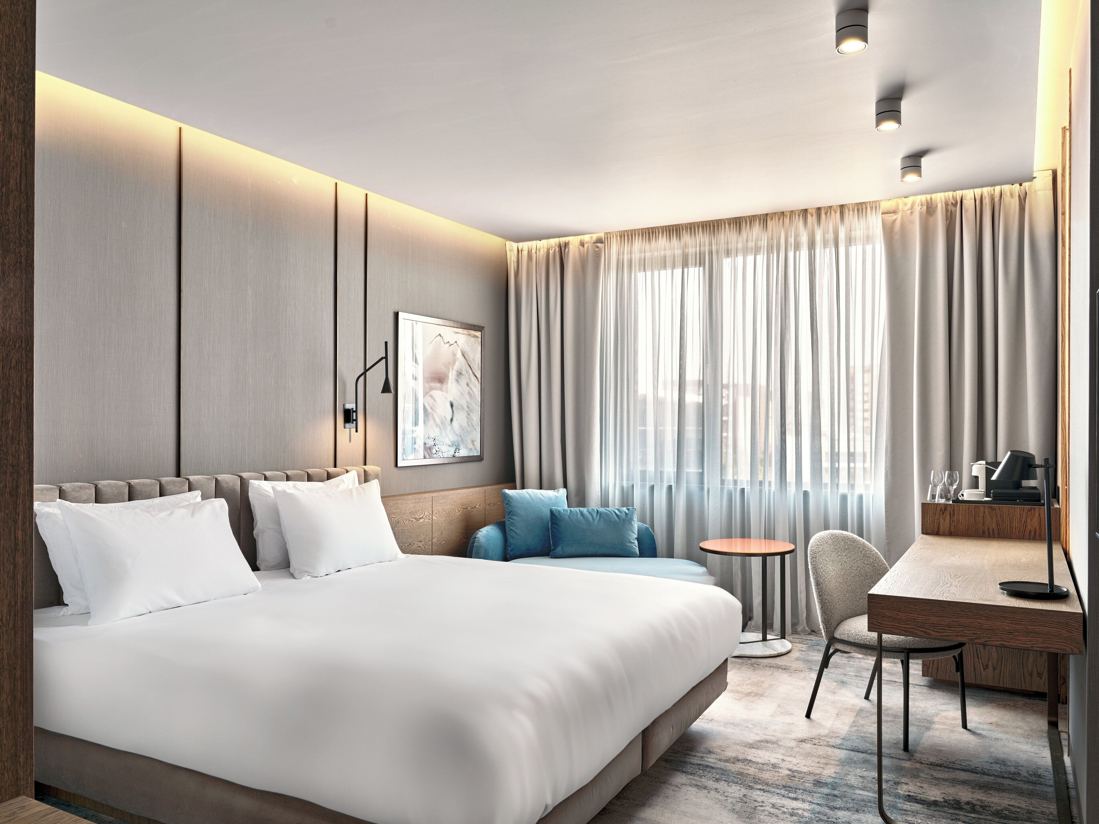 A modern hotel room with a large bed, a desk, and a chair. There is a window with curtains, a wall-mounted light, and a painting on the wall.