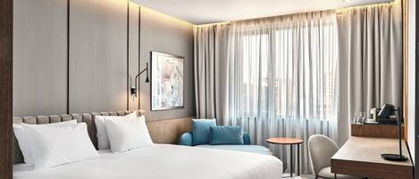 A modern hotel room with a large bed, a desk, and a chair. There is a window with curtains, a wall-mounted light, and a painting on the wall.