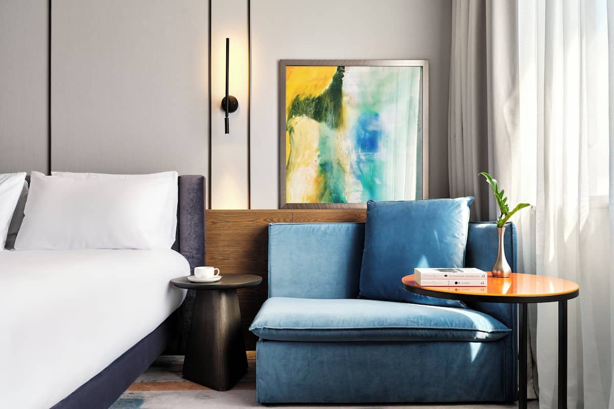 A modern hotel room with a bed, a small table with a book, a blue armchair, and a painting on the wall.