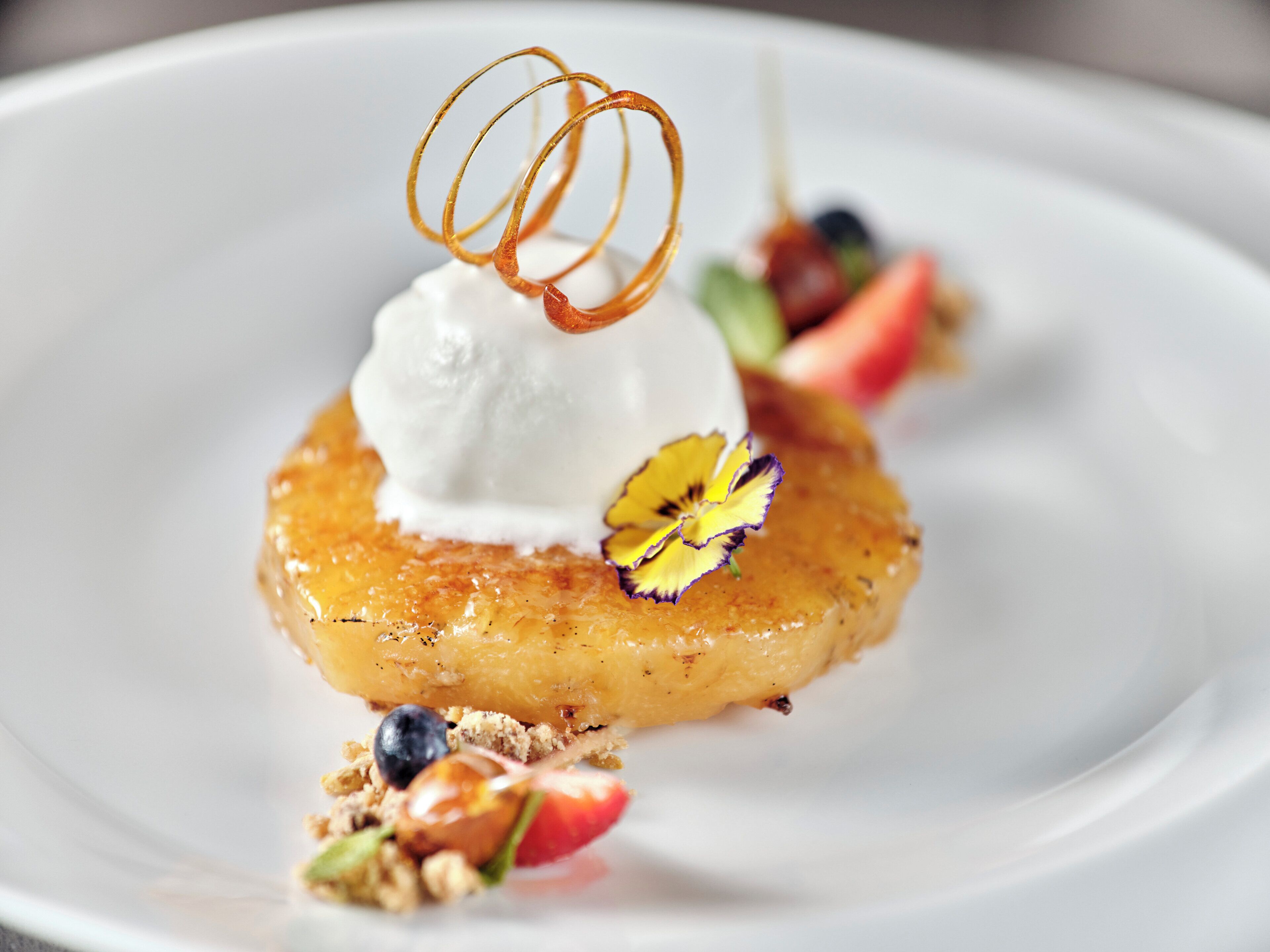 Postre sobre un plato blanco con una bola de helado, una base de fruta caramelizada y adornos que incluyen una flor y frutos rojos.