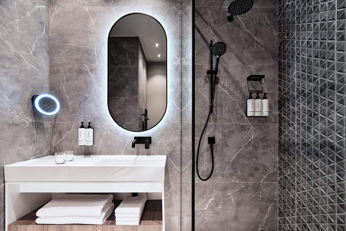 A modern bathroom with a white vanity, a large mirror, a handheld showerhead, and a wall-mounted hairdryer.