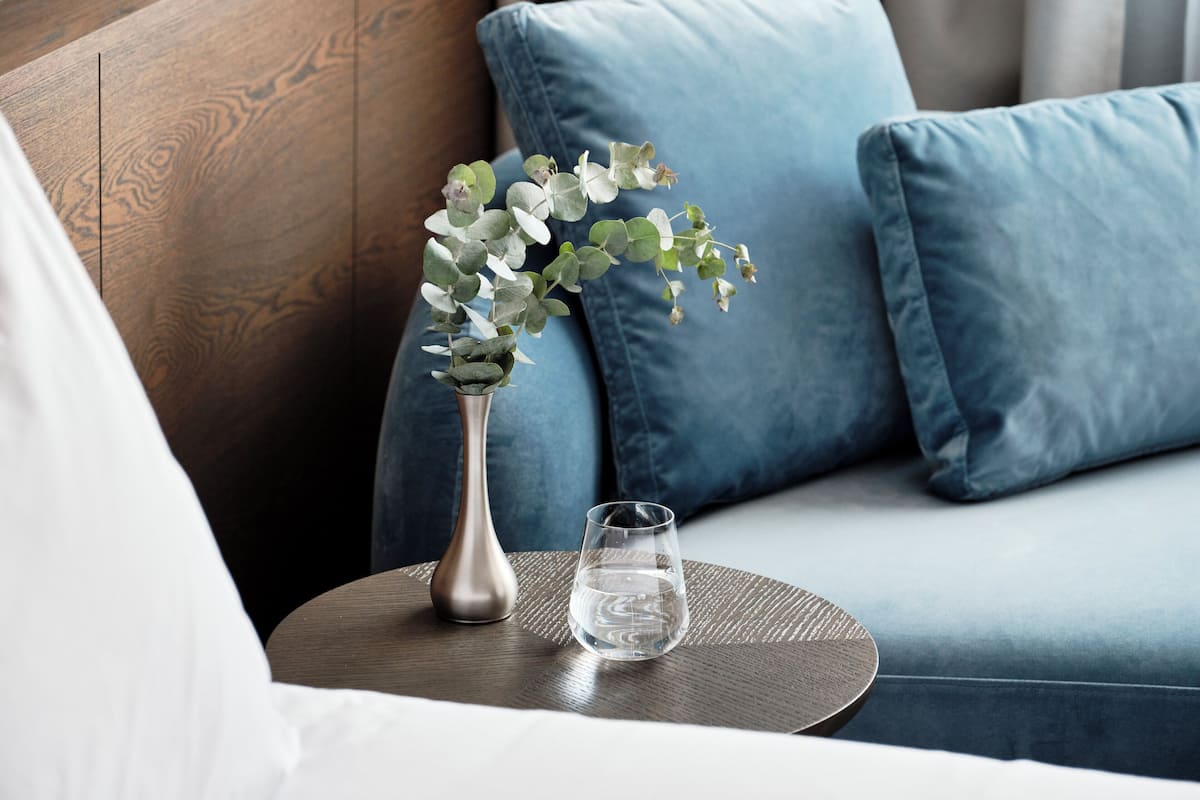 A modern living room with a blue sofa, a round wooden side table, a metallic vase with green foliage, and a clear glass of water.