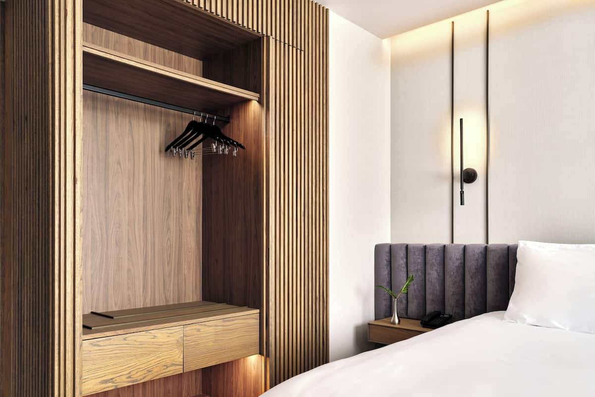A modern hotel room with a wooden wardrobe, a bedside table with a vase, and a bed with white linens.