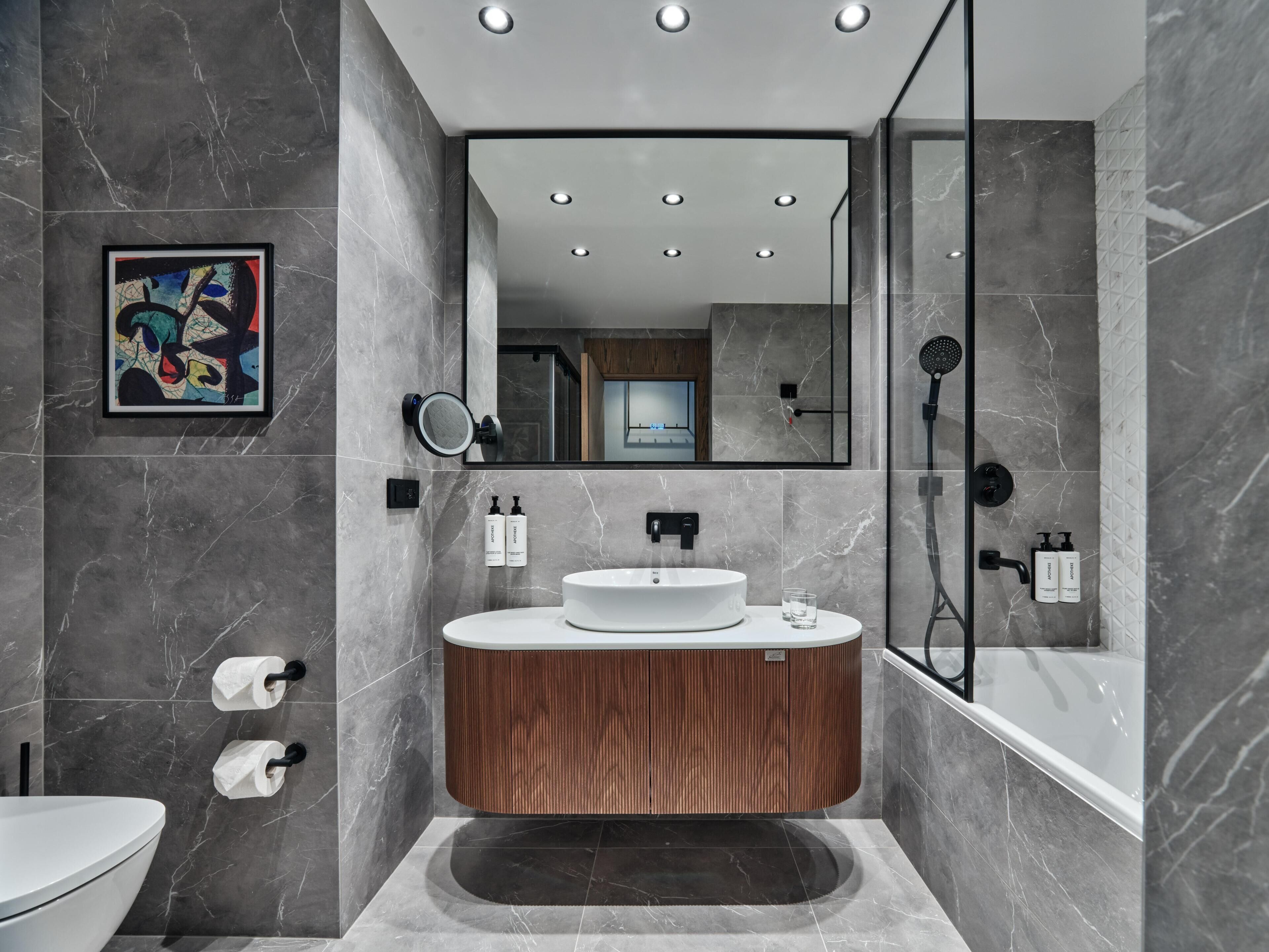 Un baño moderno con piso de mármol, tocador de madera, lavamanos blanco, espejo grande y zona de ducha.