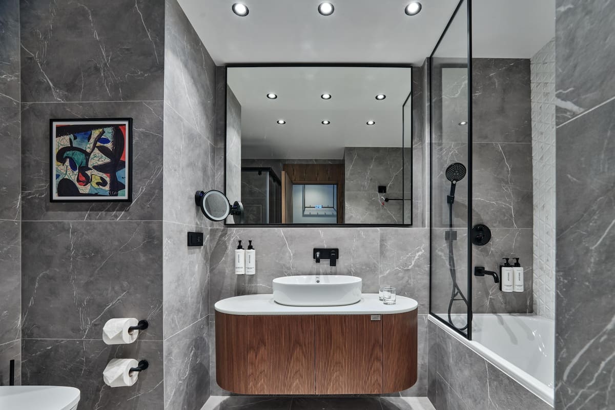 Un baño moderno con piso de mármol, tocador de madera, lavamanos blanco, espejo grande y zona de ducha.