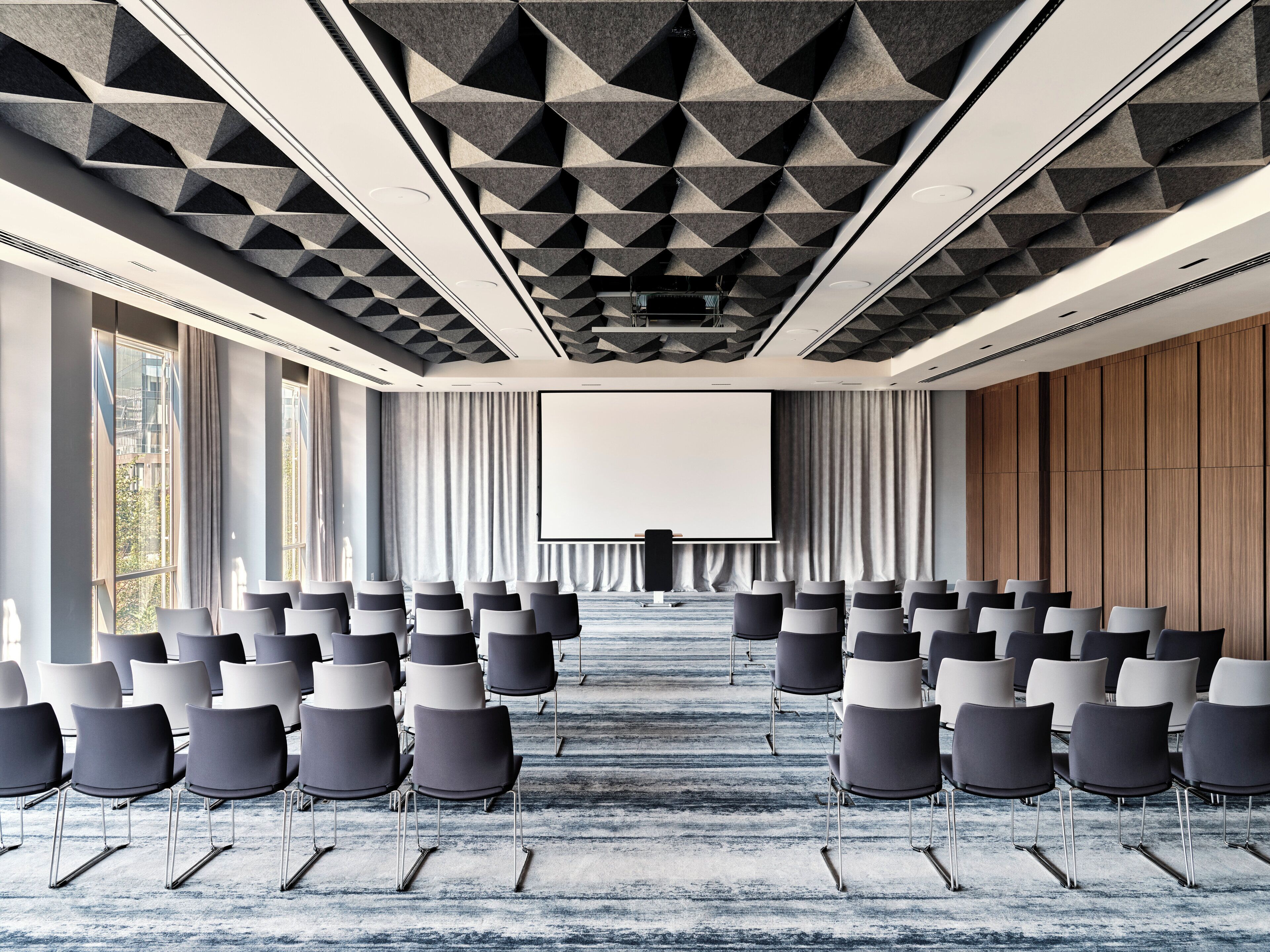 A conference room with rows of chairs facing a large screen, a patterned ceiling, and large windows.