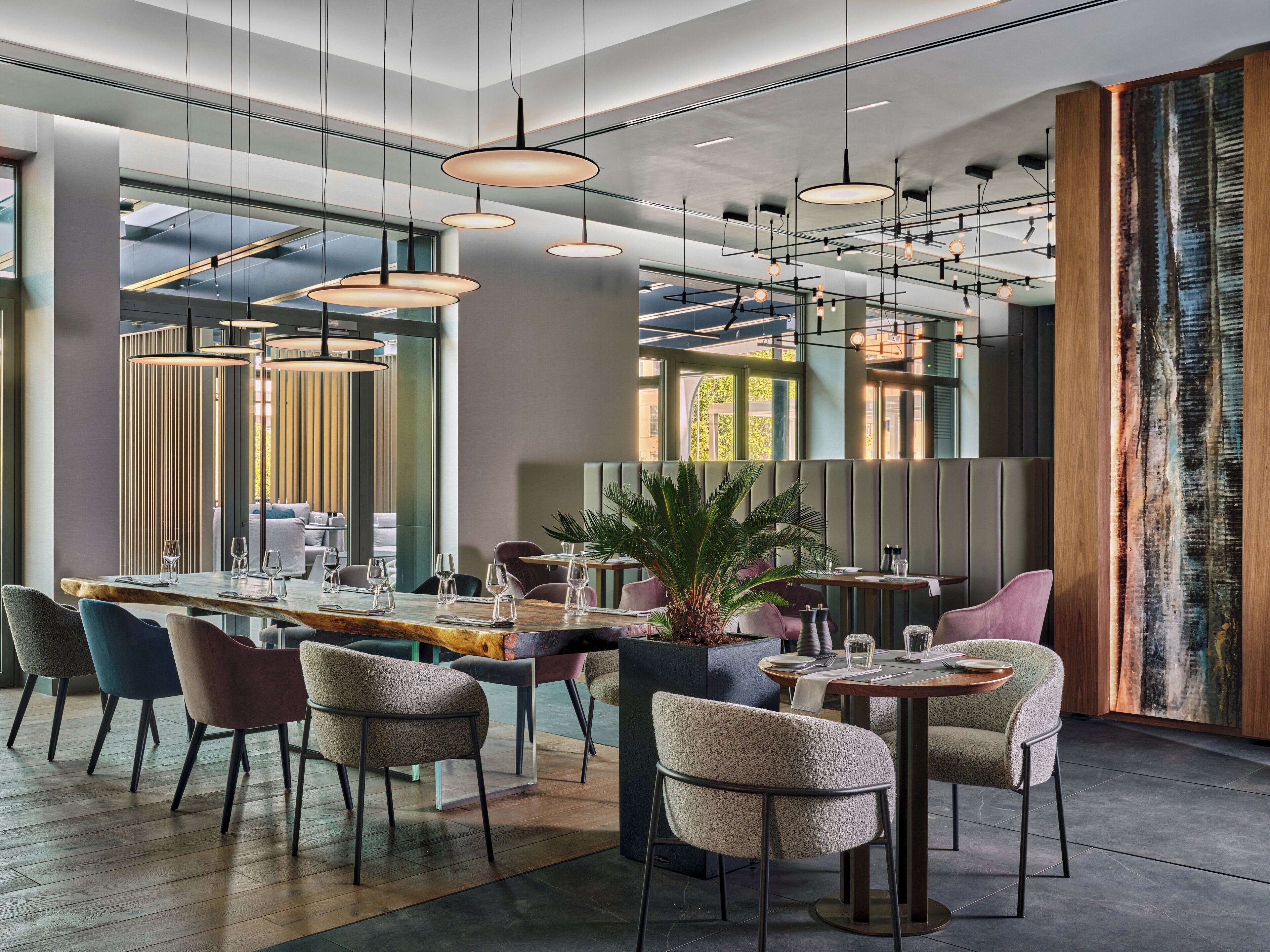 Un interior de restaurante moderno con varias mesas y sillas, iluminación colgante y un amplio ventanal que proporciona luz natural.