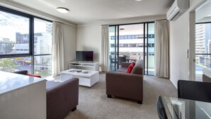 A modern hotel room with a large window, a flat-screen TV, a sofa, a glass table, and a balcony with a view of the city. - SKD SUITES (Spring Hill)