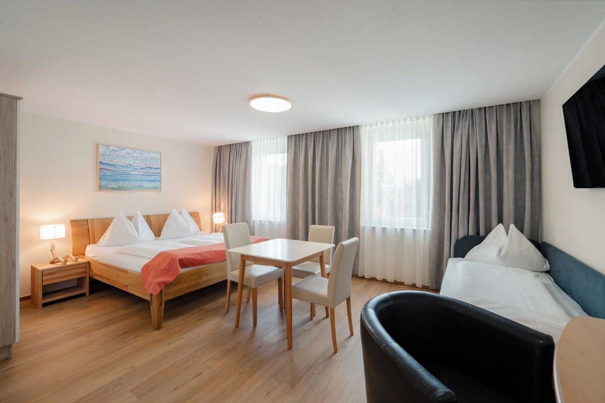 Une chambre d’hôtel comprenant un lit, un bureau, deux chaises, une petite table, une table de chevet, une lampe et un tableau au mur.