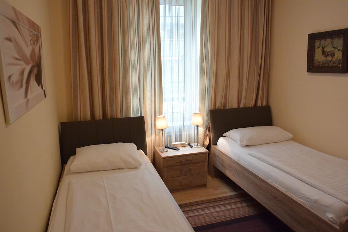 Ein Hotelzimmer mit zwei Einzelbetten, einem Nachttisch aus Holz mit Lampen, einem Fenster mit Vorhängen und einem gerahmten Bild an der Wand.