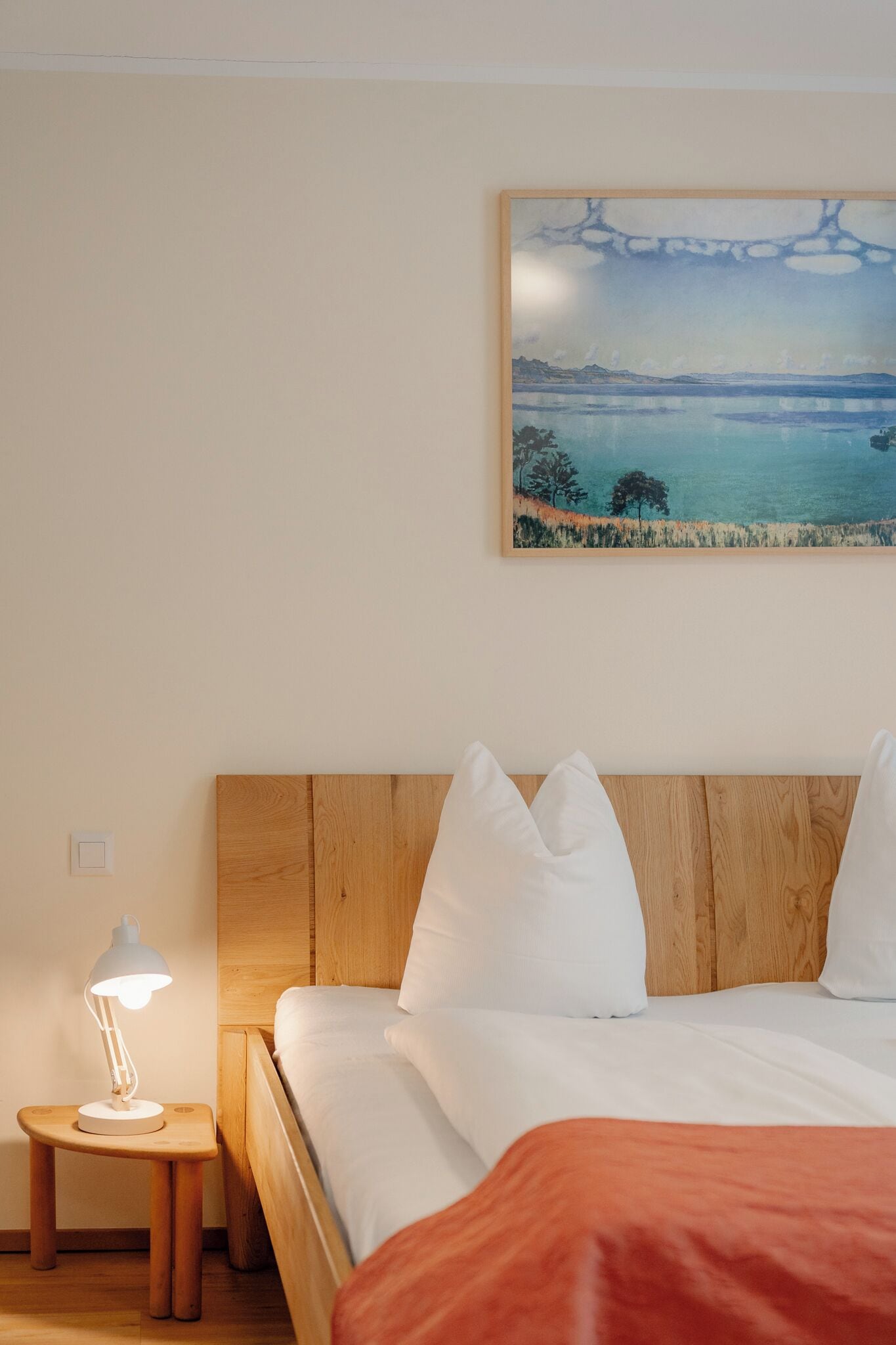A bedroom with a wooden headboard, white bedding, a nightstand with a lamp, and a painting of a landscape on the wall.