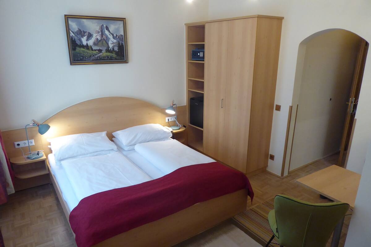Ein ordentlich bezogenes Bett mit weißen Leinen und einer roten Decke, ein Holzschrank, ein Schreibtisch mit Stuhl und ein Bergpanorama an der Wand.