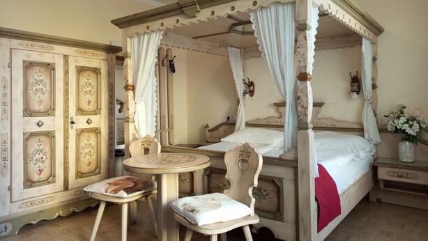 Un lit à baldaquin à quatre poteaux, avec une table et deux chaises, dans une pièce dotée de meubles richement décorés et d’un miroir.