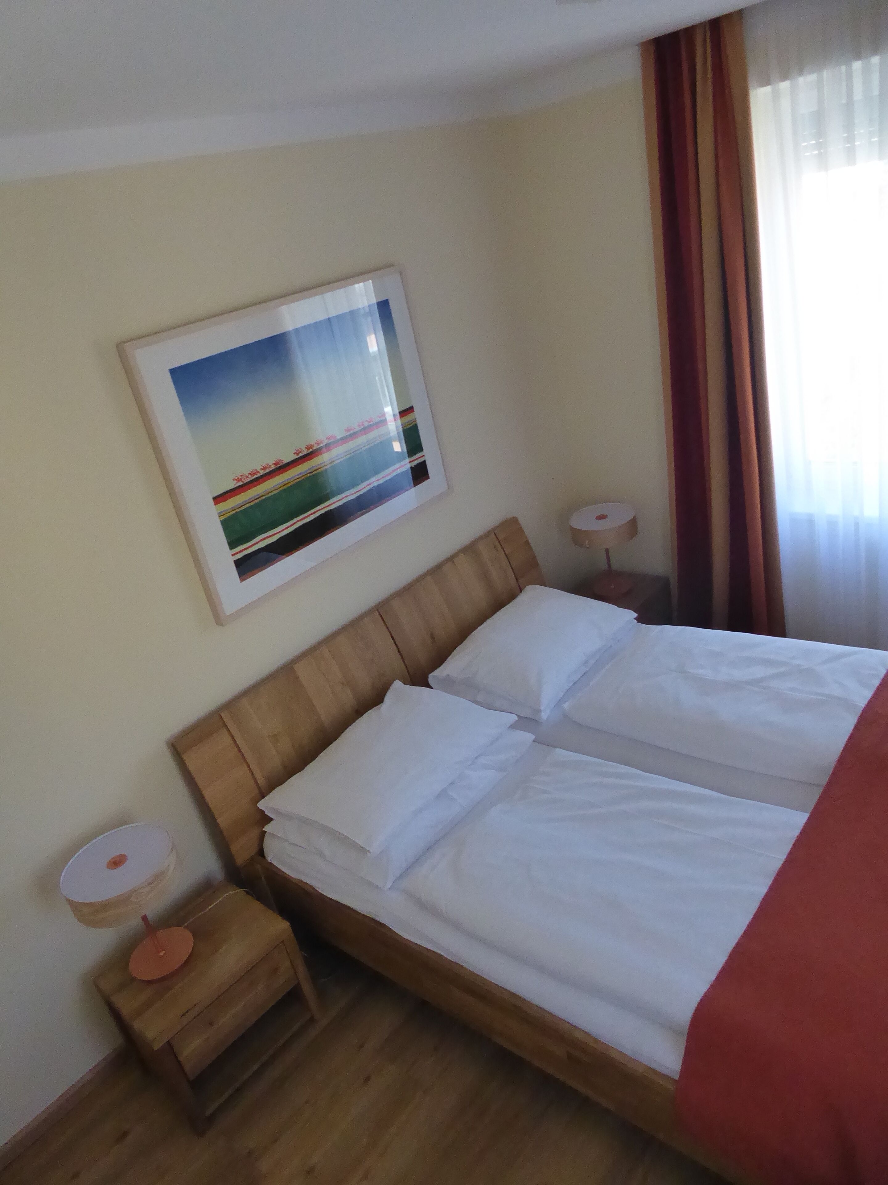 A hotel room with a wooden bed, white bedding, a framed picture on the wall, a small bedside table with a lamp, and a window with curtains.