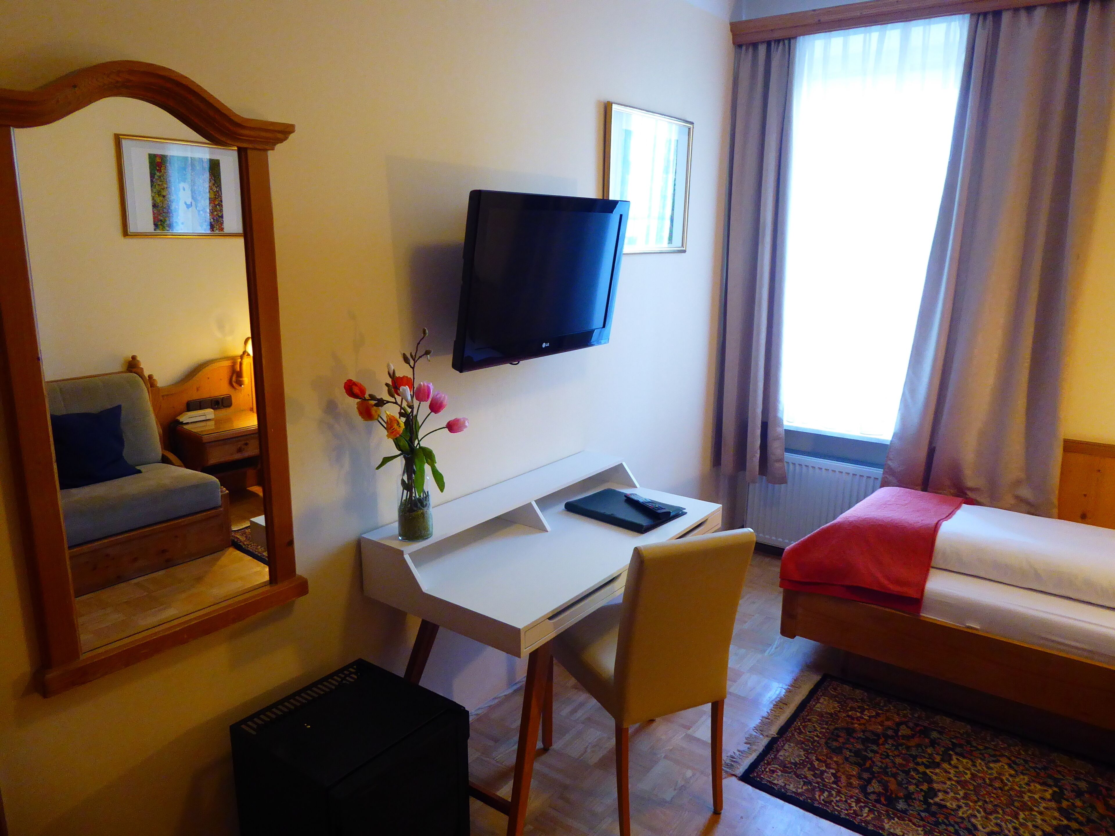 A hotel room with a bed, a desk with a chair, a television, a mirror, and a window with curtains.