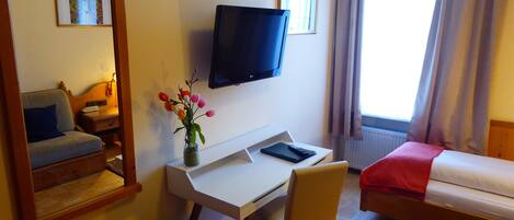 A hotel room with a bed, a desk with a chair, a television, a mirror, and a window with curtains.