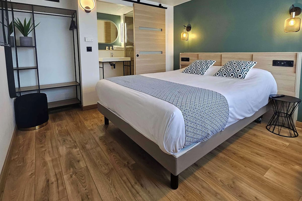 Un dormitorio moderno con una cama grande, piso de madera, baño con espejo y lavamanos, y una planta pequeña.