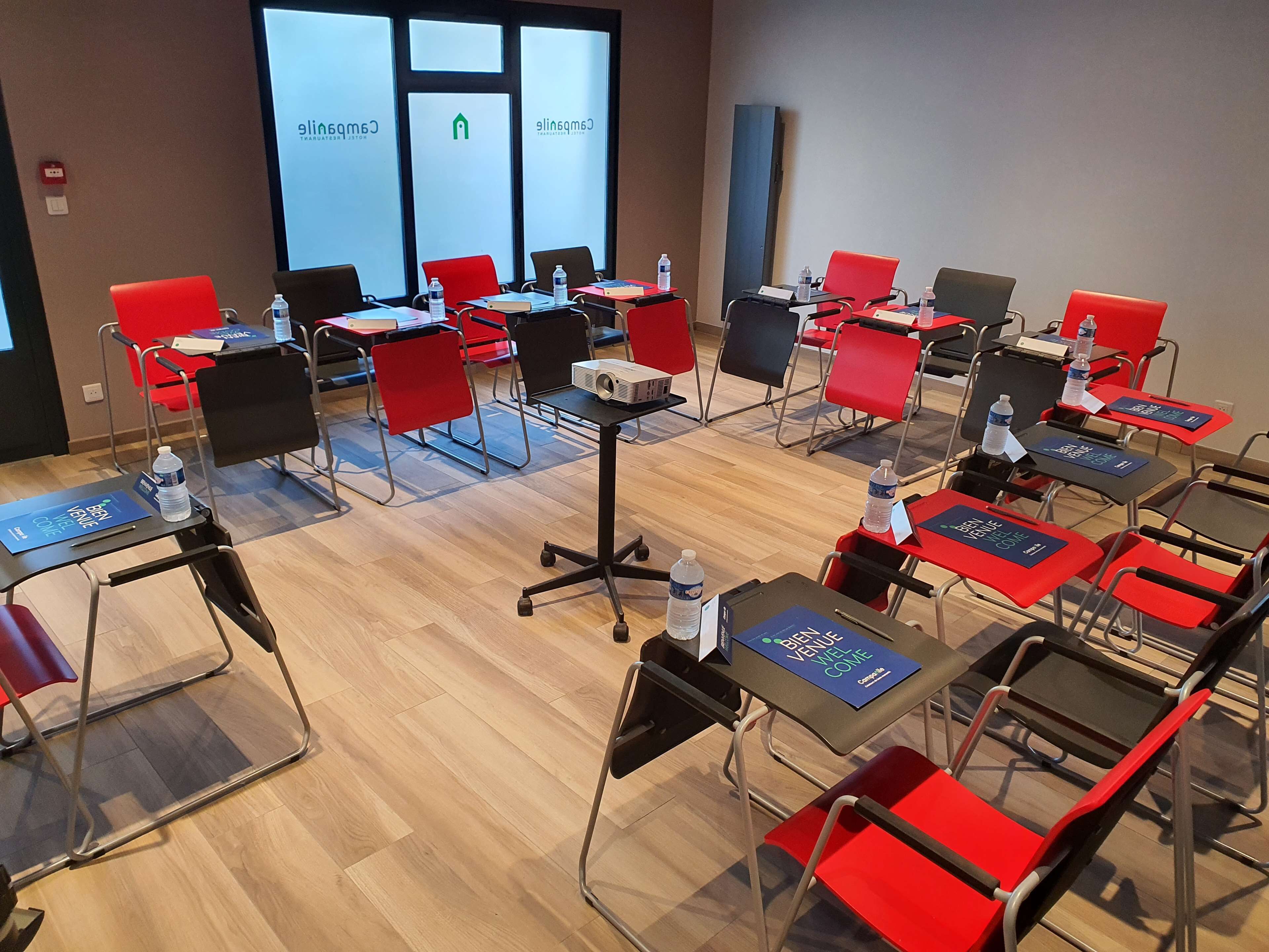 A conference room with rows of red and black chairs, tables with water bottles, and a projector on a stand.
