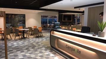 A hotel reception area with a modern design, featuring a reception desk labeled "Camparibe," seating arrangements, and a large window offering a view of the cityscape.