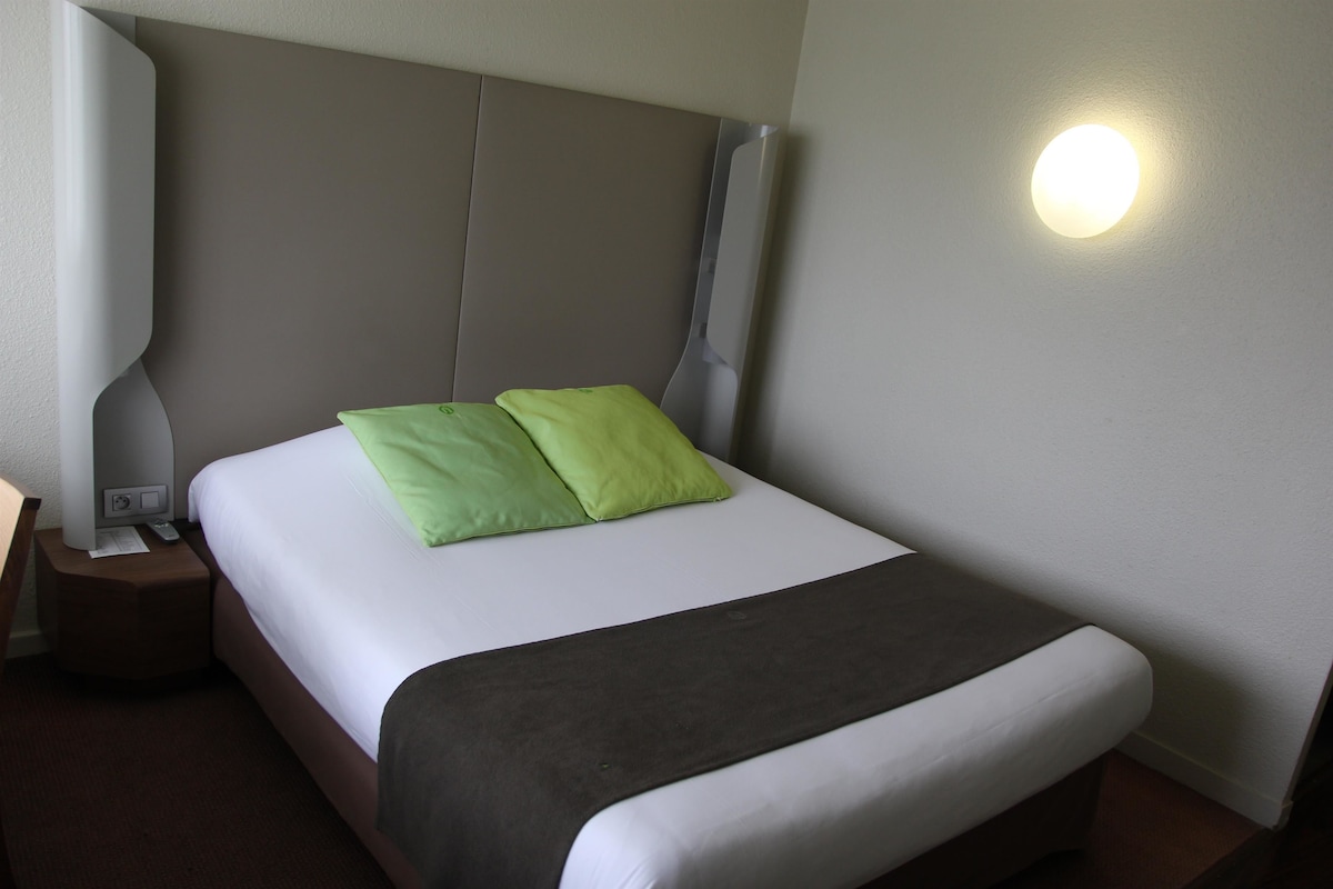 Un lit bien fait, recouvert d’une couverture blanche et grise, avec deux oreillers verts et un plaid marron.