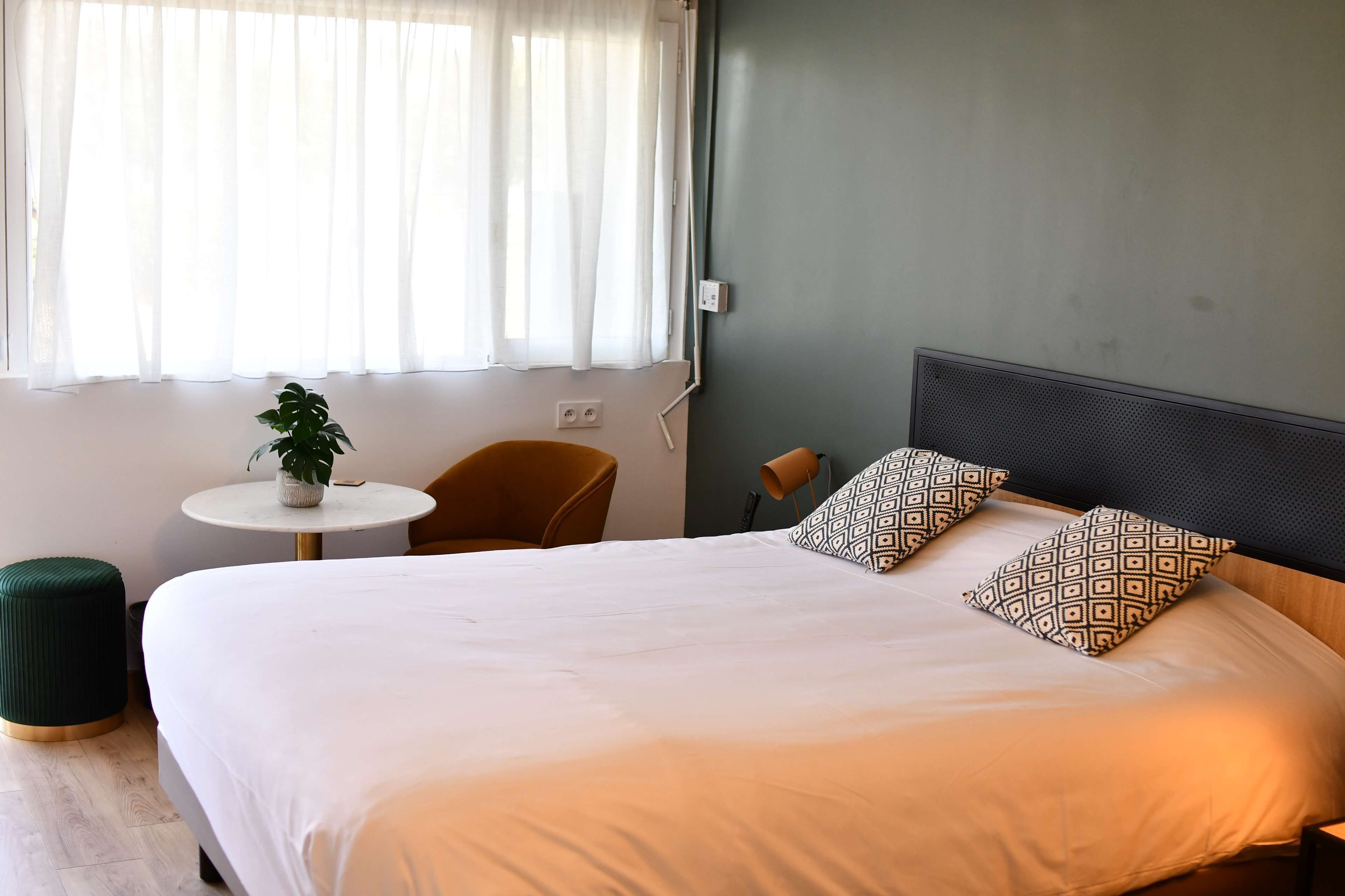 A neatly made bed with patterned pillows, a small round table with a plant, and a chair in a room with a green wall and a window with sheer curtains.