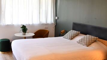 A neatly made bed with patterned pillows, a small round table with a plant, and a chair in a room with a green wall and a window with sheer curtains.