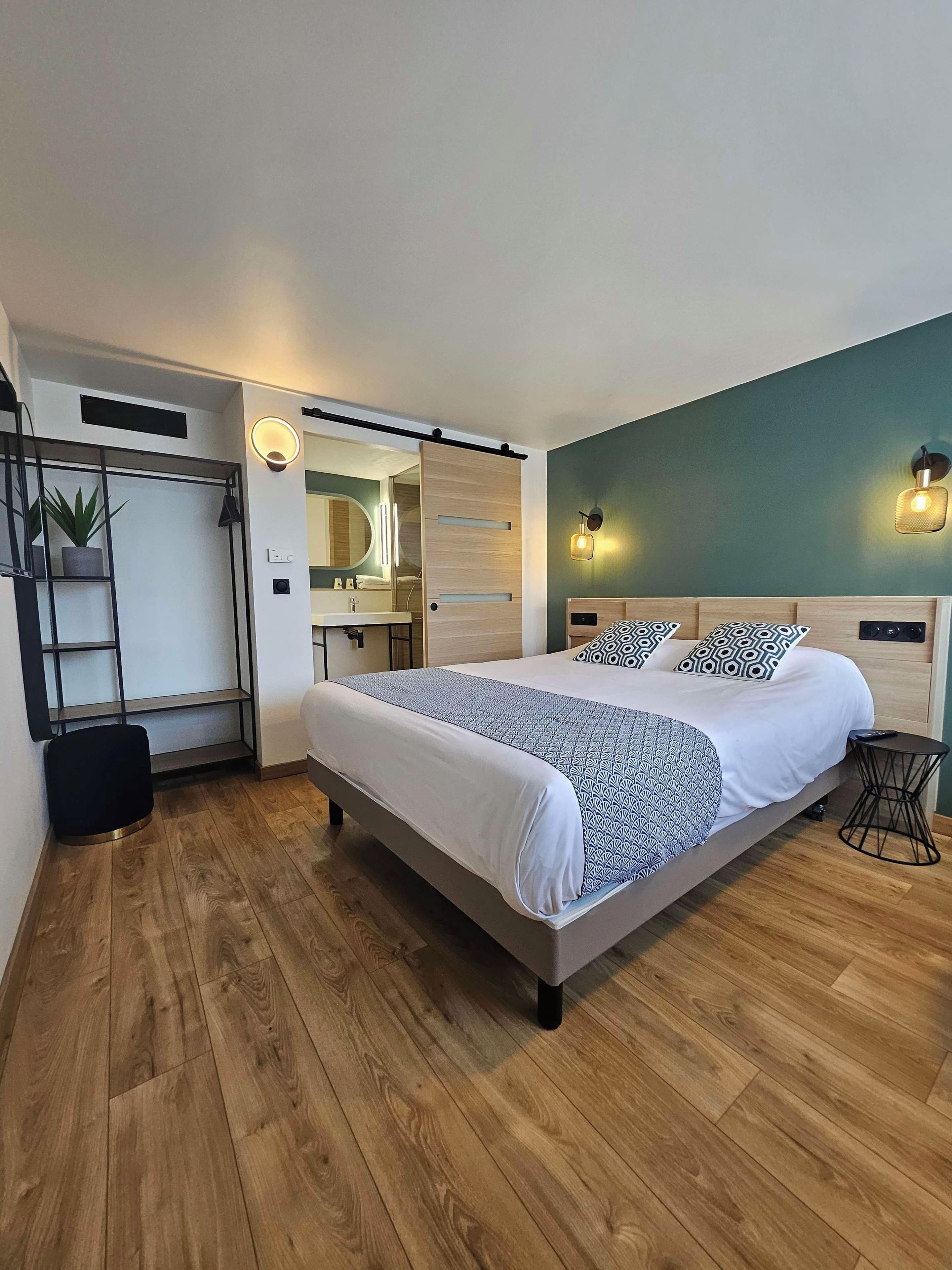 A modern bedroom with a large bed, a wooden floor, a bathroom with a mirror and sink, and a green accent wall.