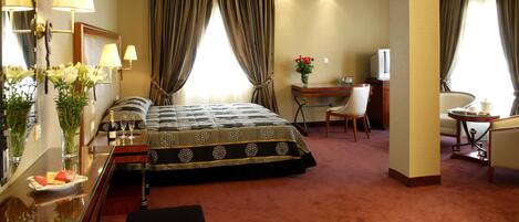 A hotel room with a large bed, a desk, a chair, a TV, and a small table with a vase of flowers.