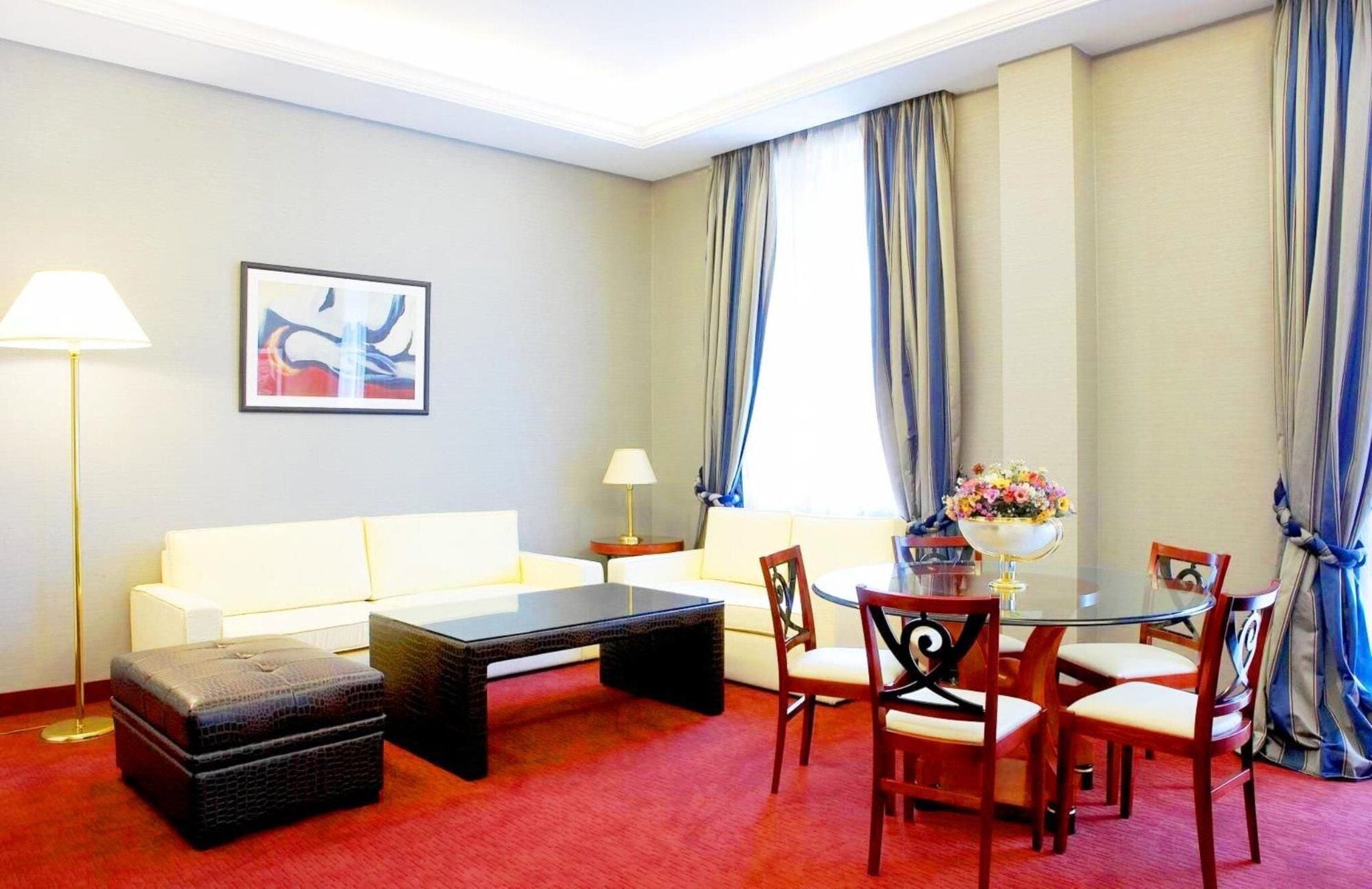 A hotel room with a sofa, a coffee table, a dining table with chairs, a lamp, and a painting on the wall.