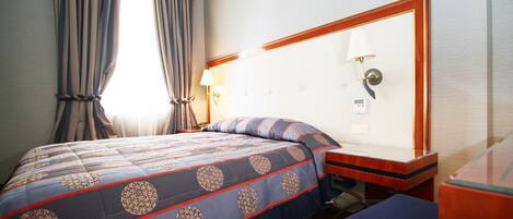 Une chambre d’hôtel avec un lit, une table de chevet, une lampe et une fenêtre avec des rideaux.