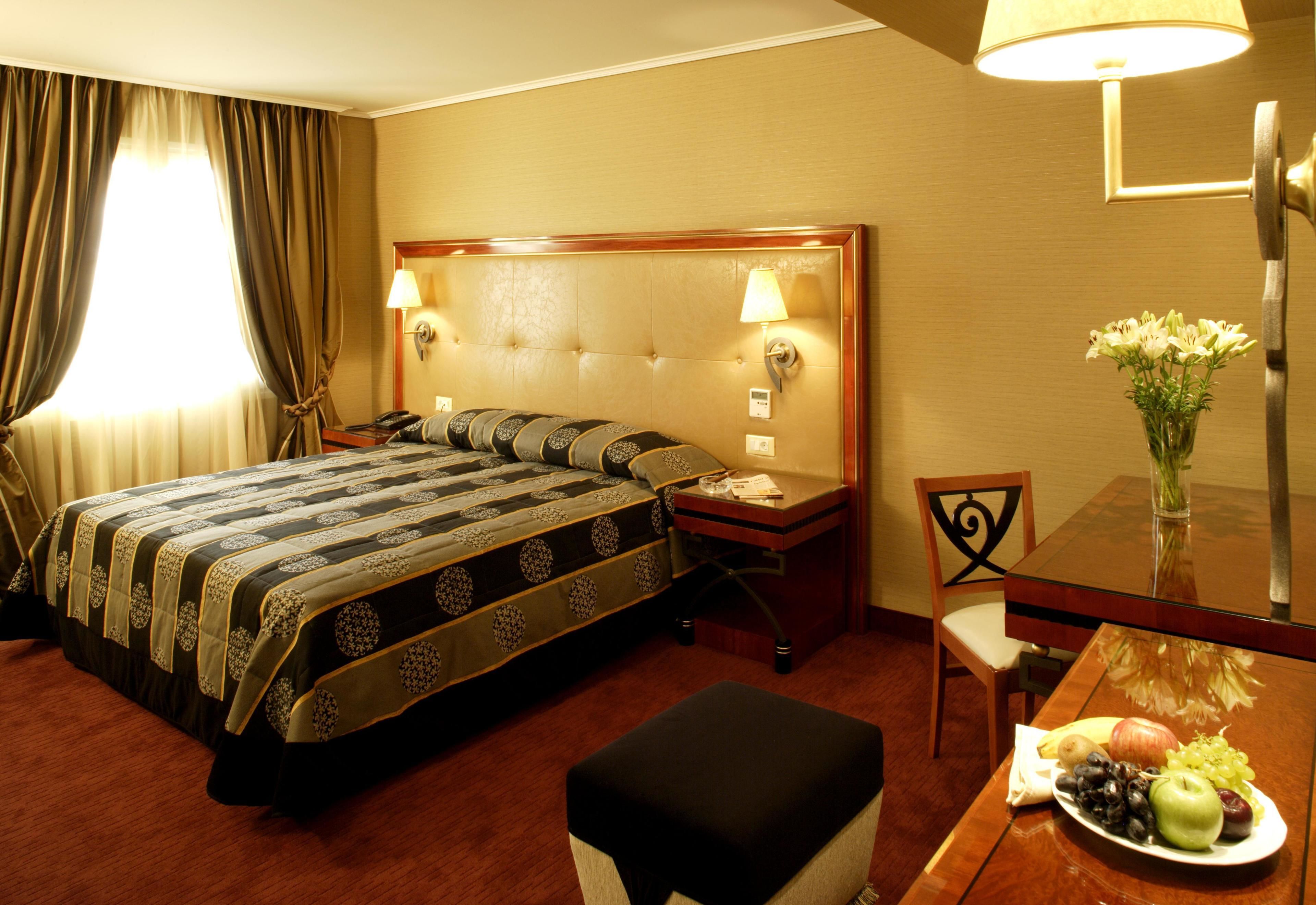 Quarto de hotel com uma cama grande, uma mesinha de cabeceira com abajur, uma pequena mesa com uma travessa de fruta e uma cadeira.