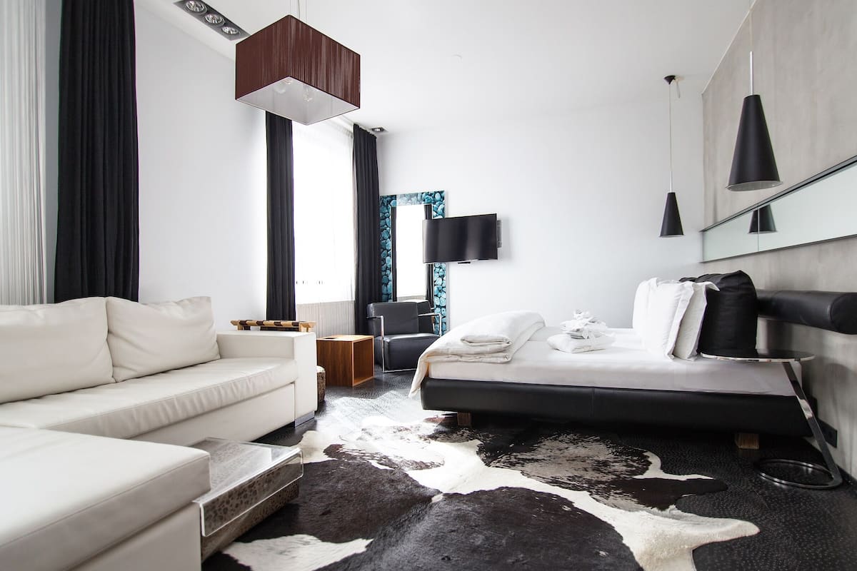 A modern hotel room with a white sofa, a bed with a black headboard, a cowhide rug, and a mounted TV.