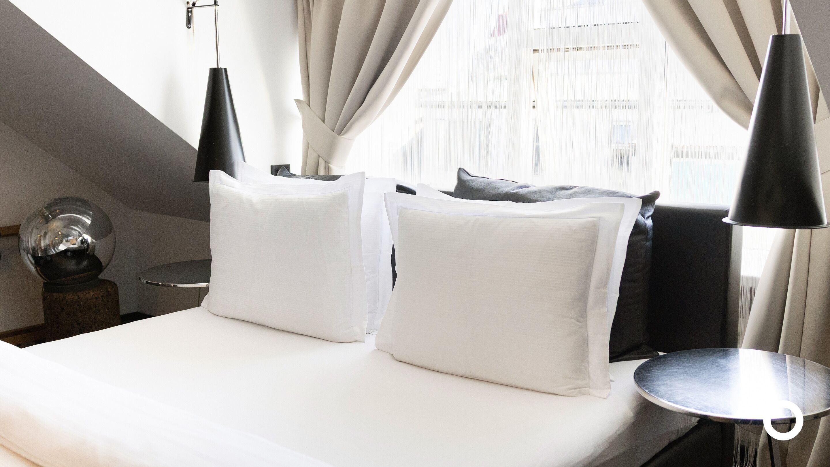 A neatly made bed with white linens and two pillows, a round side table, a modern lamp, and a decorative sphere on a stand.
