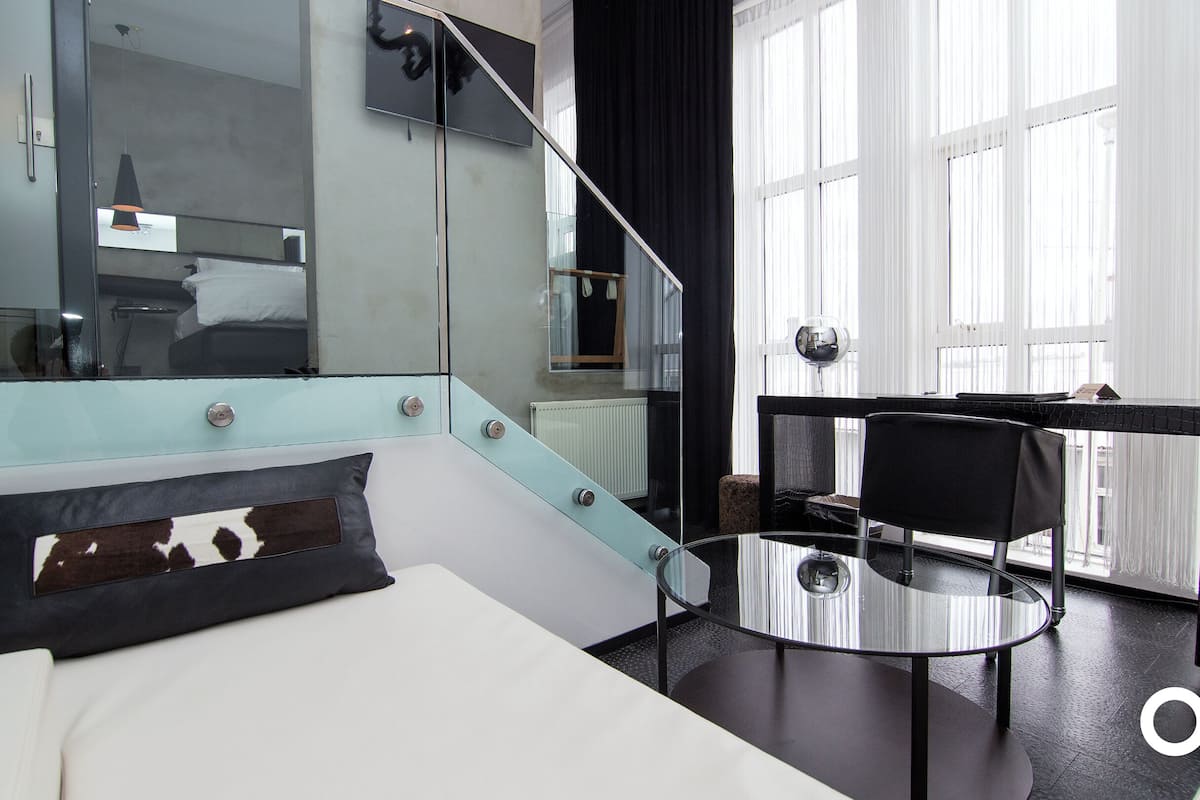 A modern hotel room with a bed, a glass table, a chair, a large mirror, and a view of the city through large windows.