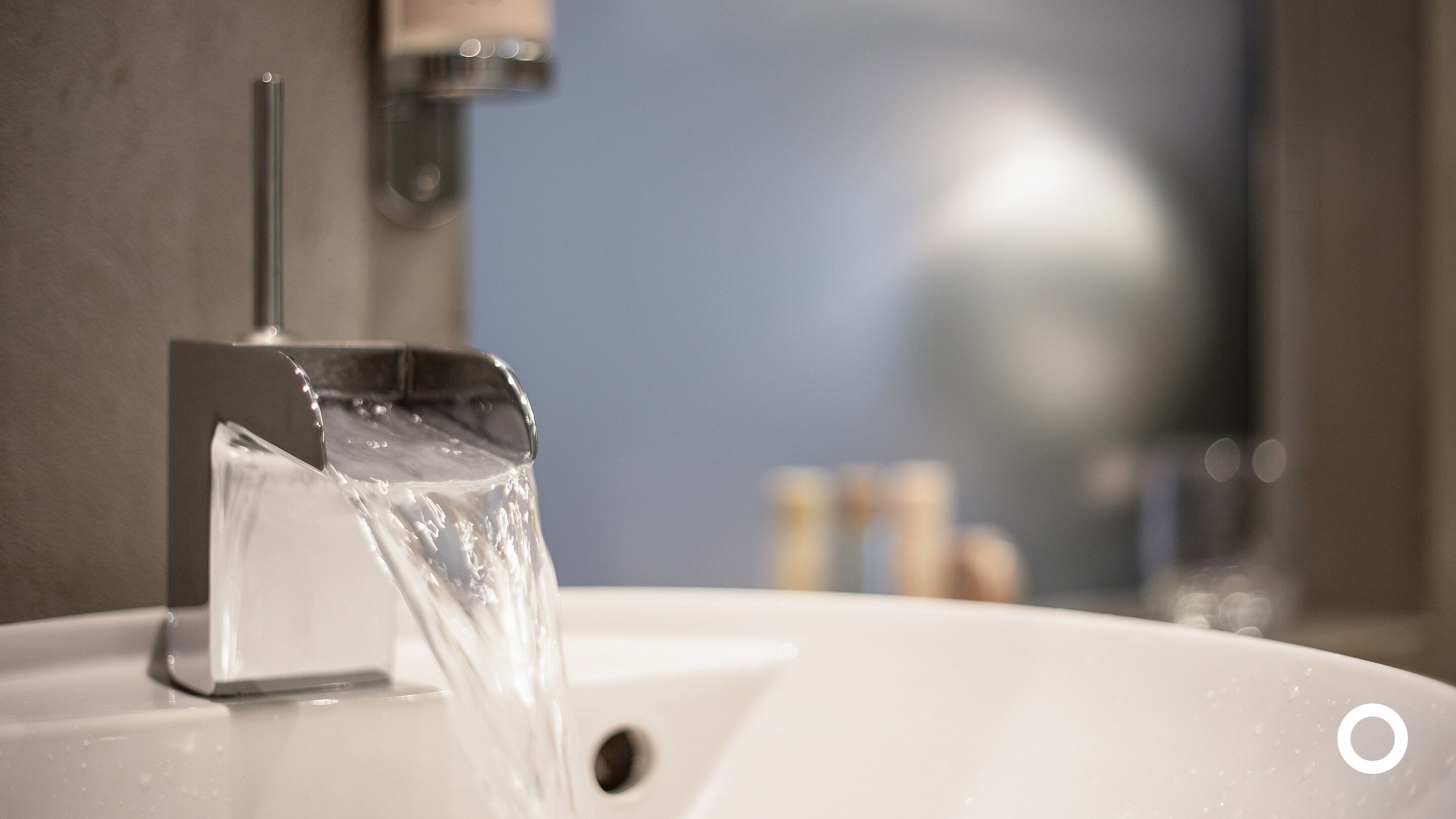 Un lavabo dans une salle de bain, avec l’eau qui coule, un miroir et des articles de toilette en arrière-plan.