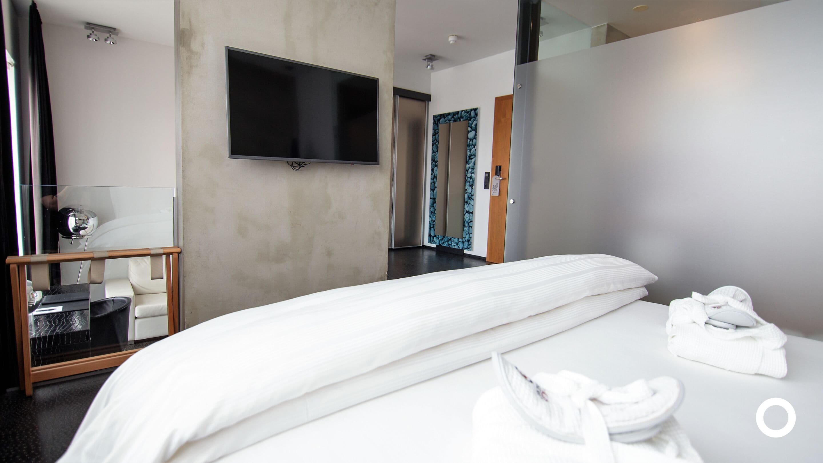 Una habitación de hotel moderna con una cama grande, un televisor de pantalla plana montado en la pared, un espejo con un marco decorativo y una mesita de noche de madera con una lámpara.