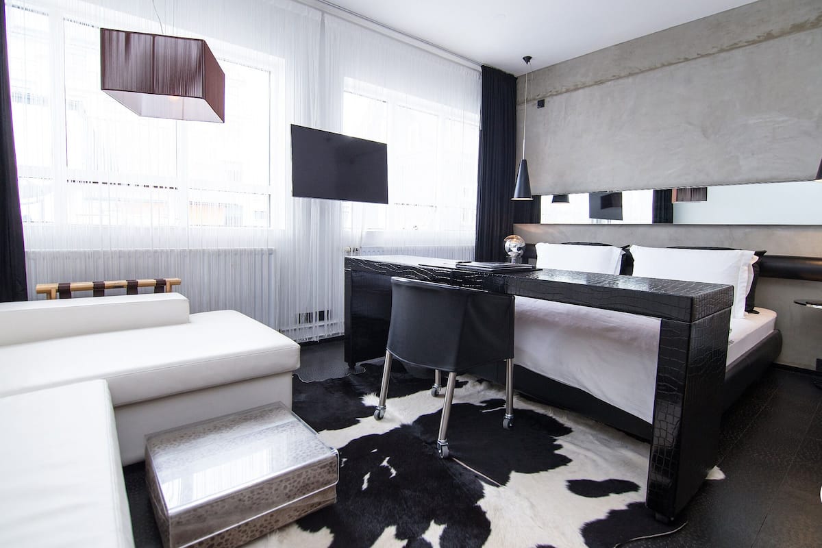 A modern hotel room with a bed, a desk, a sofa, and a large window with curtains.