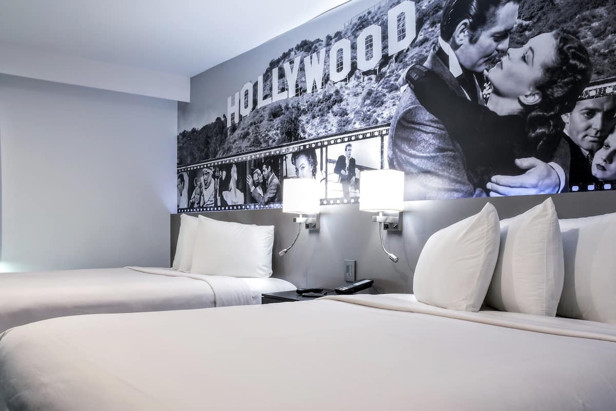 A hotel room with two beds, a large Hollywood-themed mural, and a nightstand with a lamp.