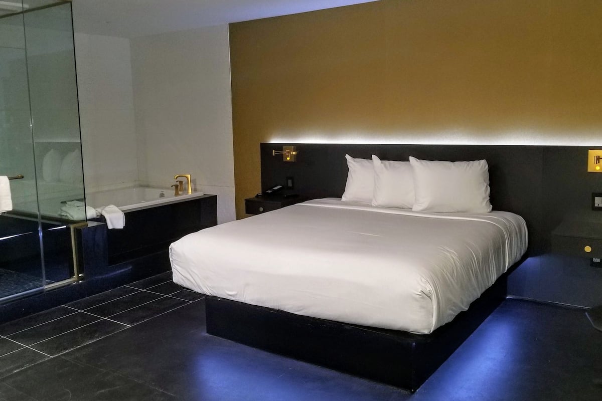 A modern hotel room with a large bed, a glass-enclosed shower, and a dark-toned nightstand.