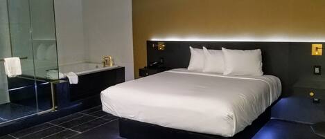 A modern hotel room with a large bed, a glass-enclosed shower, and a dark-toned nightstand.