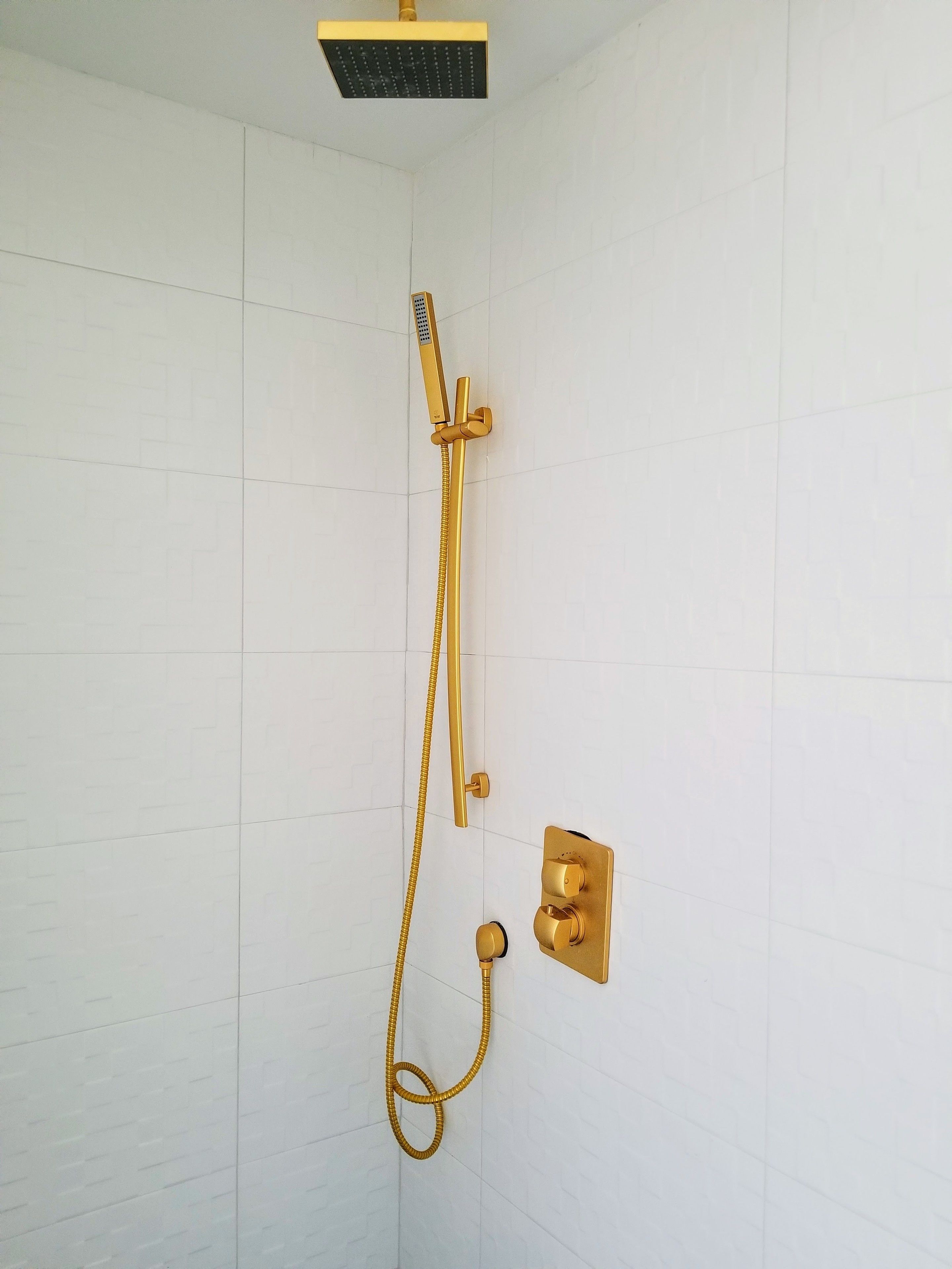 Une douche dotée d’un pommeau et d’un flexible dorés, fixés sur un mur recouvert de carreaux blancs.